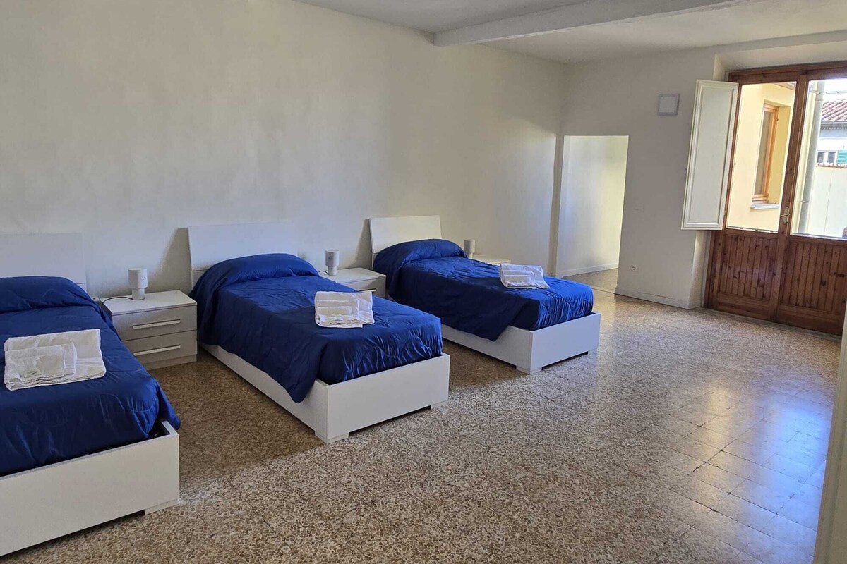 Une chambre avec trois lits, chacun doté d’une tête de lit blanche et de draps bleus. Il y a une table de chevet blanche avec une lampe sur chaque lit. La chambre a un sol carrelé et une porte en bois.