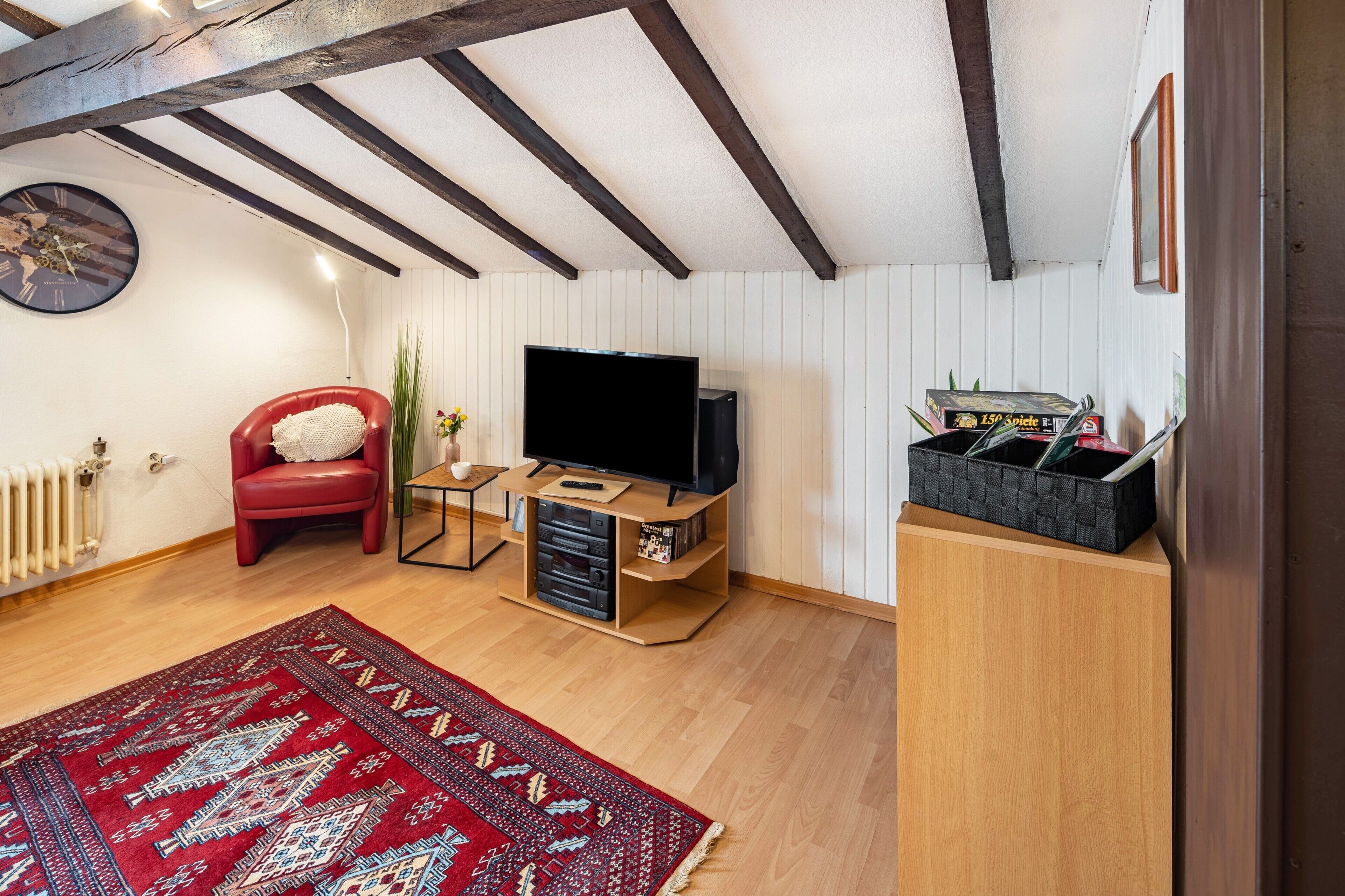A room with a wooden floor, a red chair, a television, a bookshelf, and a radiator.