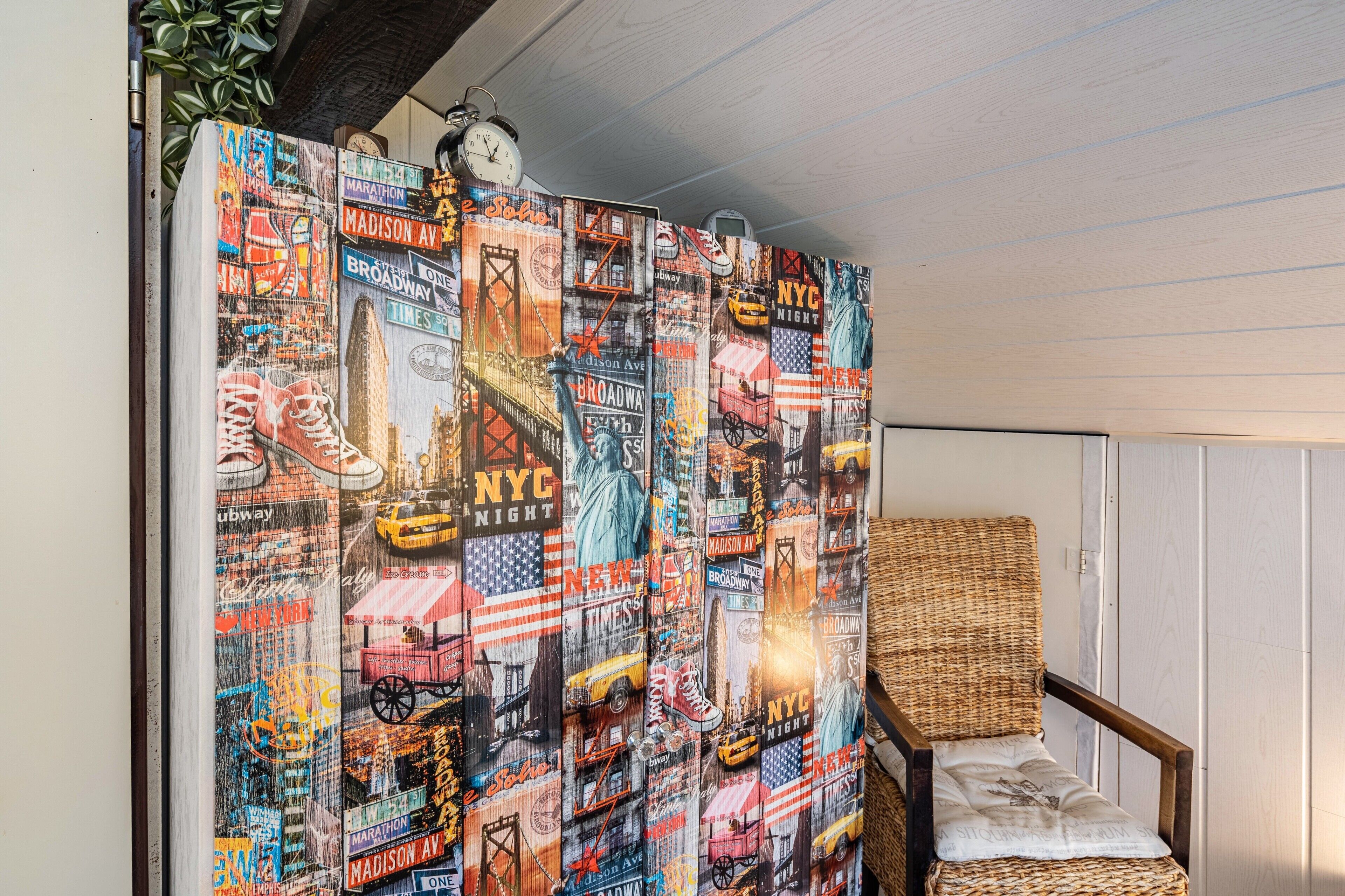 Een kamer met een groot, kleurrijk gordijn met een stadsgezicht, een rieten stoel en een klok aan de muur.