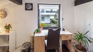 Een kamer met een houten bureau, een stoel, een plant in pot, een klok en een raam met uitzicht op een gebouw.