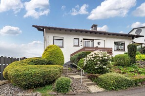 Ein zweistöckiges Haus mit weißer Fassade, braunem Dach und einer kleinen Veranda mit Stufen zum Eingang.