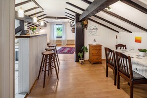 Ein Zimmer mit Holzbalkendecke, einem Esstisch mit Stühlen, einer Bar mit Hockern und Blick ins Freie.