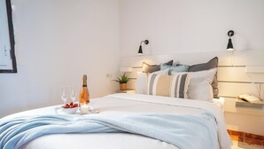 Ein ordentlich bezogenes Bett mit weißer und hellblauer Bettdecke, ein Nachttisch mit Lampe, ein kleines Topfpflänzchen sowie eine Champagnerflasche mit Gläsern und Erdbeeren auf dem Nachttisch.
