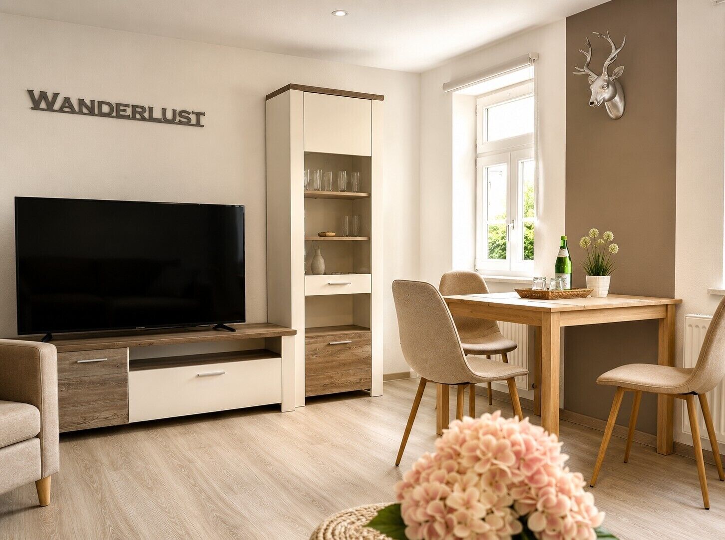 A modern living room with a flat-screen TV, a bookshelf, a dining table with chairs, and a decorative deer head on the wall.