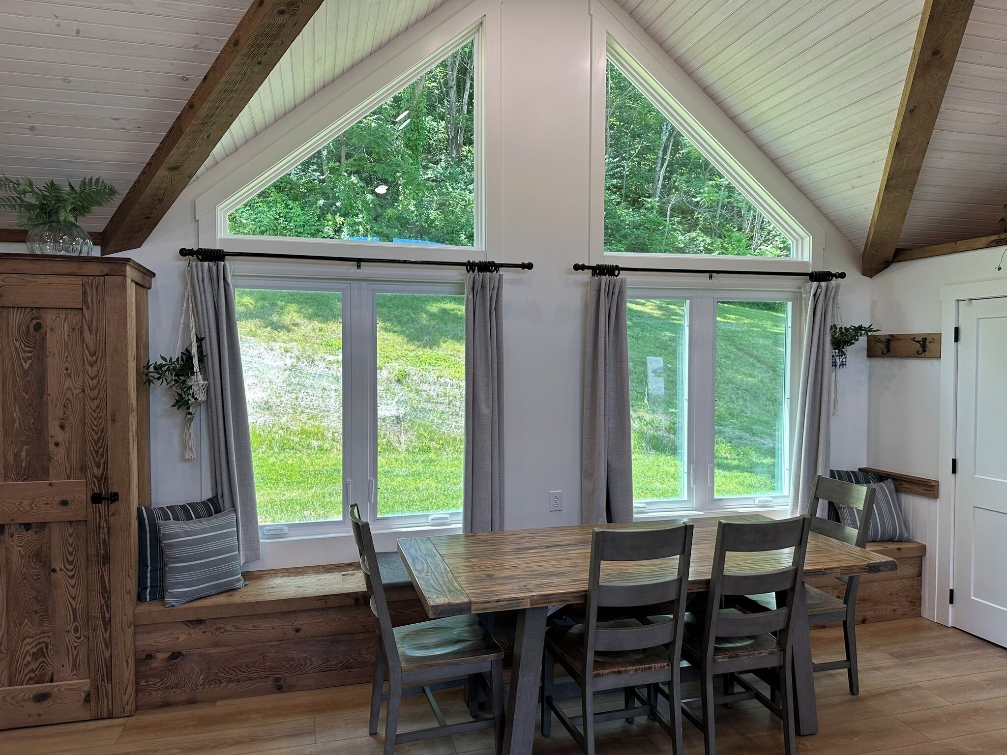 Un espace repas avec une table et des chaises en bois, de grandes fenêtres avec des rideaux et une vue sur la verdure à l’extérieur.