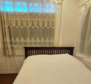 Una cama con cabecera de madera, una ventana con cortinas traslúcidas y suelo de madera.