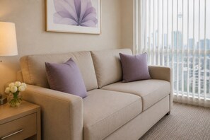Ein beiges Sofa mit zwei violetten Kissen, eine Vase mit weißen Blumen und ein gerahmtes Bild an der Wand.