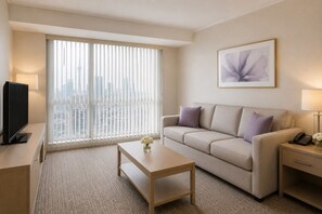 Ein Hotelzimmer mit Sofa, Couchtisch, Fernseher, Telefon und Fenster mit Jalousien.