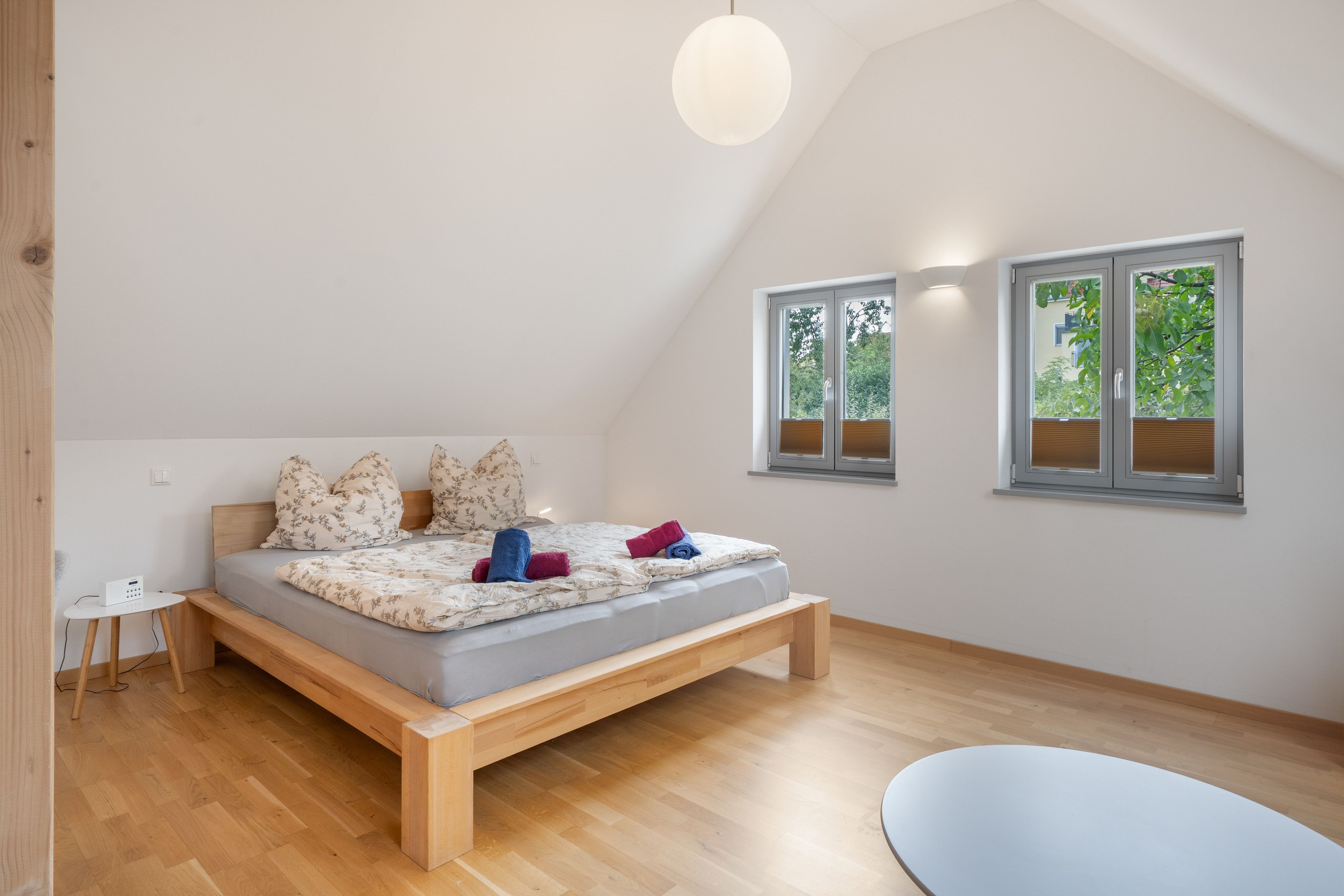 A modern bedroom with a sloped ceiling, a wooden bed frame, a bedside table, and two windows with blinds.