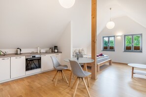 A modern kitchen with a built-in oven, a dining area with a round table and chairs, and a view of greenery through the windows. - Apartment 'Ziegfried#Ziegenhof' (Radebeul)