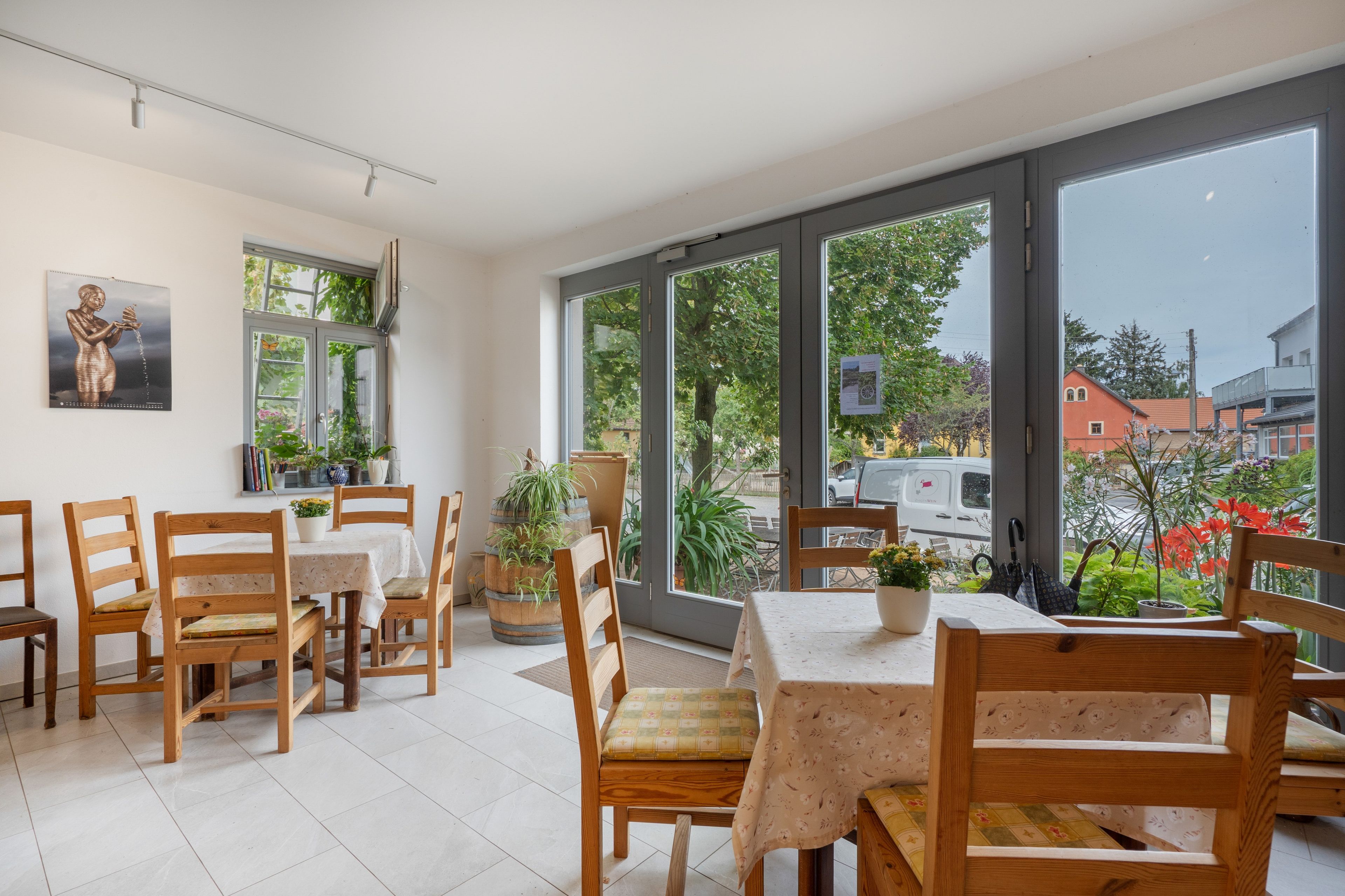 A dining area with wooden chairs and tables, large windows with a view of the outside, and a wall-mounted picture.