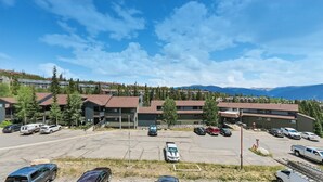 En parkeringsplats med flera bilar, en byggnad med ett sluttande tak och en bergig bakgrund.
