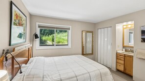 Ein Schlafzimmer mit Bett, einem Fenster mit Blick ins Grüne, einer Nachttischlampe, einem Spiegel und einem Badezimmer mit Waschbecken und Handtuch.