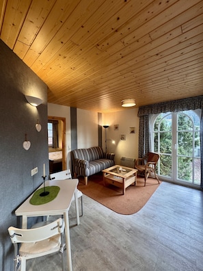 A cozy living room with a wooden ceiling, a sofa, a coffee table, a dining table with chairs, and a large window with a view of trees.