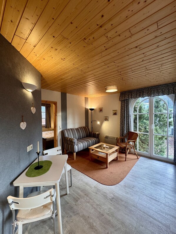 Un salon chaleureux avec un plafond en bois, un canapé, une table basse, une table à manger avec des chaises et une grande fenêtre donnant sur la forêt.