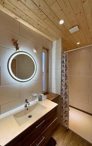 Une salle de bain avec un plafond en bois, un miroir rond avec un éclairage intégré, un lavabo avec un robinet et un rideau de douche.