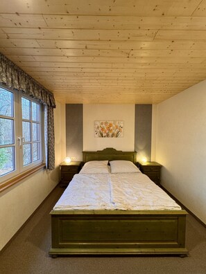 Une chambre avec un plafond en bois, un lit avec un cadre vert, deux tables de chevet avec des lampes, une fenêtre avec des rideaux et une œuvre d’art accrochée au mur.