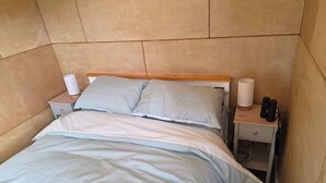 Una cama con cabecera de madera, dos almohadas, una mesita de noche con una lámpara y un cajón pequeño.