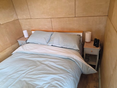 A bed with a wooden headboard, two pillows, a nightstand with a lamp, and a small drawer.. Cosy Welsh Glamping Escape at Bumblebee Lodge