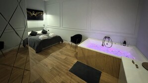 A modern bedroom with a bed, a black chair, a wooden floor, a white bathtub with a light, and a ceiling fan. - Apartament z Jacuzzi Kraków GREY Miodowa (Kraków)