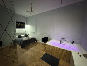 Ein modernes Schlafzimmer mit Bett, schwarzem Sessel, Holzboden, weißer Badewanne mit Lampe und Deckenventilator.
