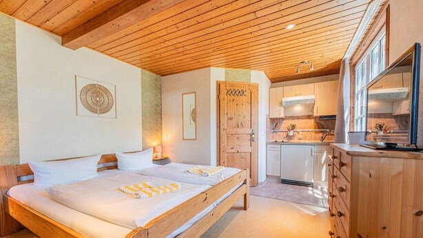 Ein Schlafzimmer mit zwei Betten, einer Holzbalkendecke, einer Küche mit weißen Schränken und einer Holzkommode.