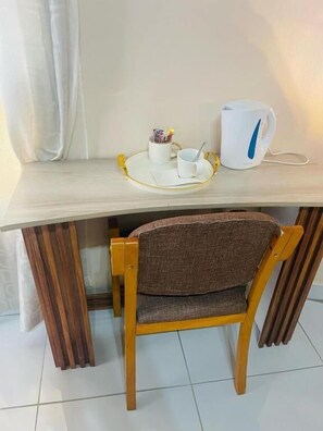 Ein Holzstuhl mit gewebter Sitzfläche, eine weiße Tasse, eine weitere weiße Tasse und ein weißer Wasserkocher auf einem Holztisch.