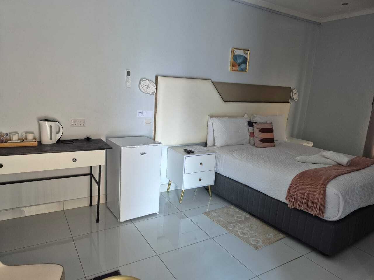 A modern hotel room with a bed, bedside table, a small white refrigerator, a desk with a chair, and a coffee maker.