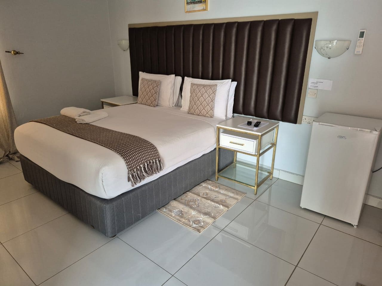 Una habitación de hotel moderna con una cama grande, mesitas de noche, un refrigerador y una lámpara fijada a la pared.