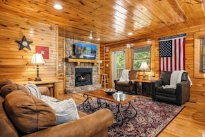 A cozy log cabin interior with a stone fireplace, a television mounted above it, and an American flag. - New Listing | 5BR Gatlinburg Cabin w/ Hot Tub + Game Room + Free Tickets (Gatlinburg)