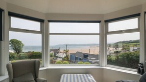 A room with a bay window overlooking a beach, a grey armchair, a striped ottoman, and a black sofa. - Hillside (Tyn-y-Gongl)
