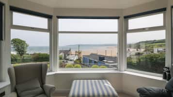 A room with a bay window overlooking a beach, a grey armchair, a striped ottoman, and a black sofa.