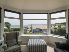 A room with a bay window overlooking a beach, a grey armchair, a striped ottoman, and a black sofa. - Hillside (Tyn-y-Gongl)
