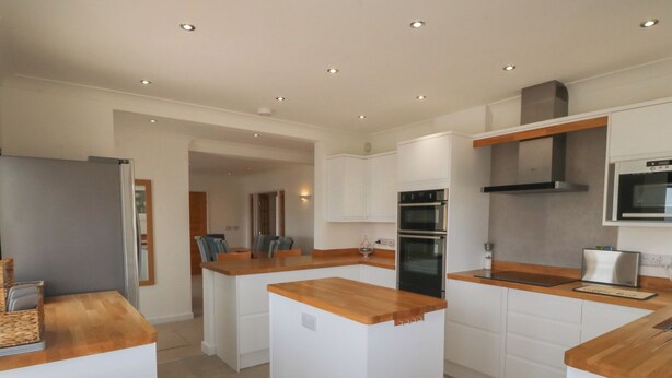 A modern kitchen with white cabinetry, wooden countertops, and a central island with seating. - Hillside (Tyn-y-Gongl)
