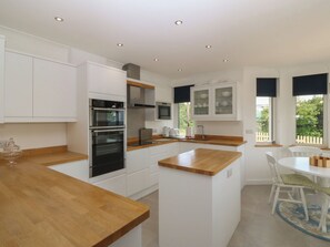 A modern kitchen with white cabinetry, a wooden countertop, built-in ovens, a microwave, and a dining area with a white table and chairs. - Hillside (Tyn-y-Gongl)