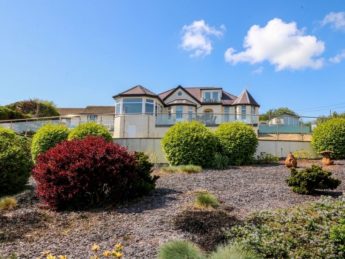 A large house with a balcony, surrounded by well-maintained gardens and a gravel driveway.