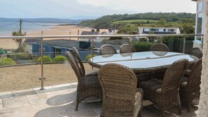 A balcony with wicker furniture, a glass table, and a view of a beach and hills. - Hillside (Tyn-y-Gongl)