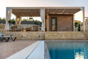 Ein Poolbereich mit Steinmauer, Holzterrasse und einer überdachten Sitzgelegenheit mit Kissen und Tisch.