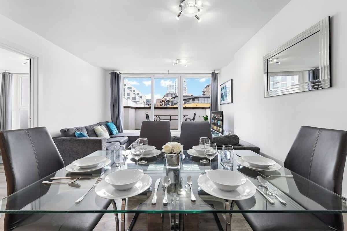 A modern dining area with a glass table set for four, surrounded by black chairs, with a city view through large windows.