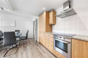 A modern kitchen with wooden cabinets, a built-in oven, and a dining area with a table and chairs. - Zone 2 Serviced apartment by the River Thames (London)