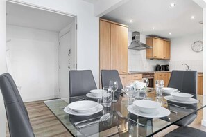 A modern kitchen with wooden cabinets, a stainless steel range hood, and a dining area with a glass table set for four. - Zone 2 Serviced apartment by the River Thames (London)