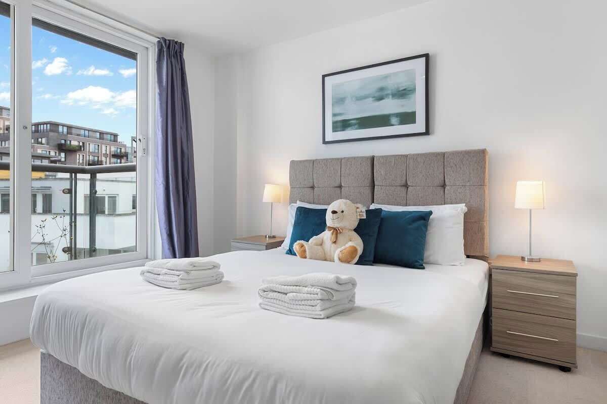 Habitación de hotel con una cama grande, dos mesitas de noche, una ventana con cortinas, un cuadro enmarcado en la pared y un peluche sobre la cama.
