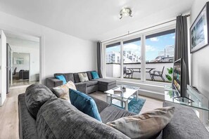 Uma sala de estar moderna com um sofá em formato de L, uma mesa de centro de vidro e uma janela grande com vista para prédios e céu.