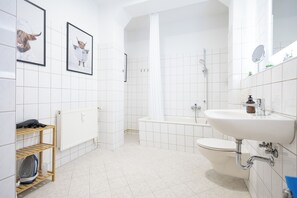 Un baño moderno con lavamanos, inodoro y ducha blancos. Hay cuadros enmarcados en la pared, un estante de madera con una tetera y un radiador.