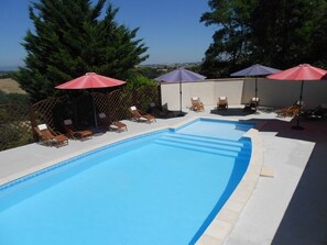 Ein Swimmingpool mit Liegestühlen und Sonnenschirmen, umgeben von Bäumen und blauem Himmel.