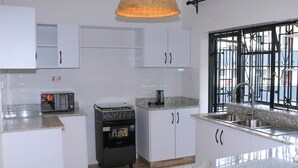Una cocina con armarios blancos, microondas, estufa, cafetera, un amplio ventanal con reja y una lámpara colgante.