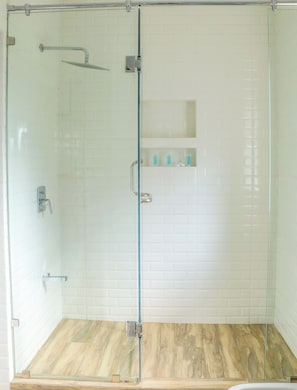 A glass-enclosed shower with a wooden floor, a metal showerhead, and a shelf with toiletries. - Rose Crown Suites (Nairobi)