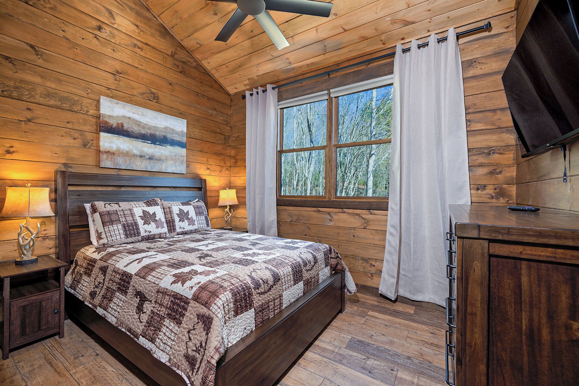 Un dormitorio tipo cabaña de madera con una cama, dos lámparas de noche, un ventilador de techo, una ventana con cortinas y un televisor en la pared.