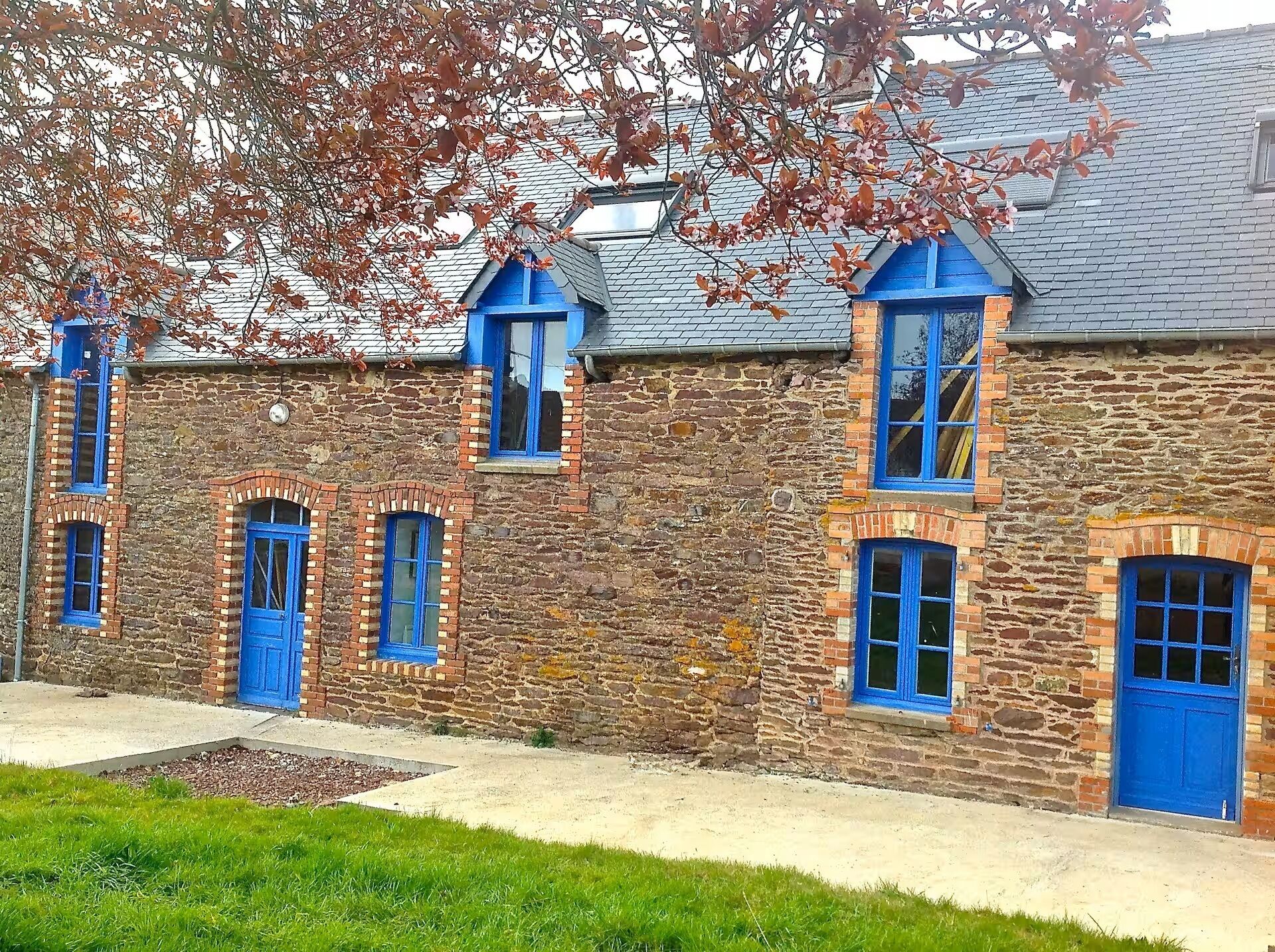 Un edificio de piedra con puertas y ventanas azules, techo inclinado y un pequeño jardín delantero.