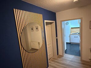Ein Flur mit blauer Wand, einer Holzlamellenverkleidung, einer weißen Tür und Blick in einen Küchenbereich.